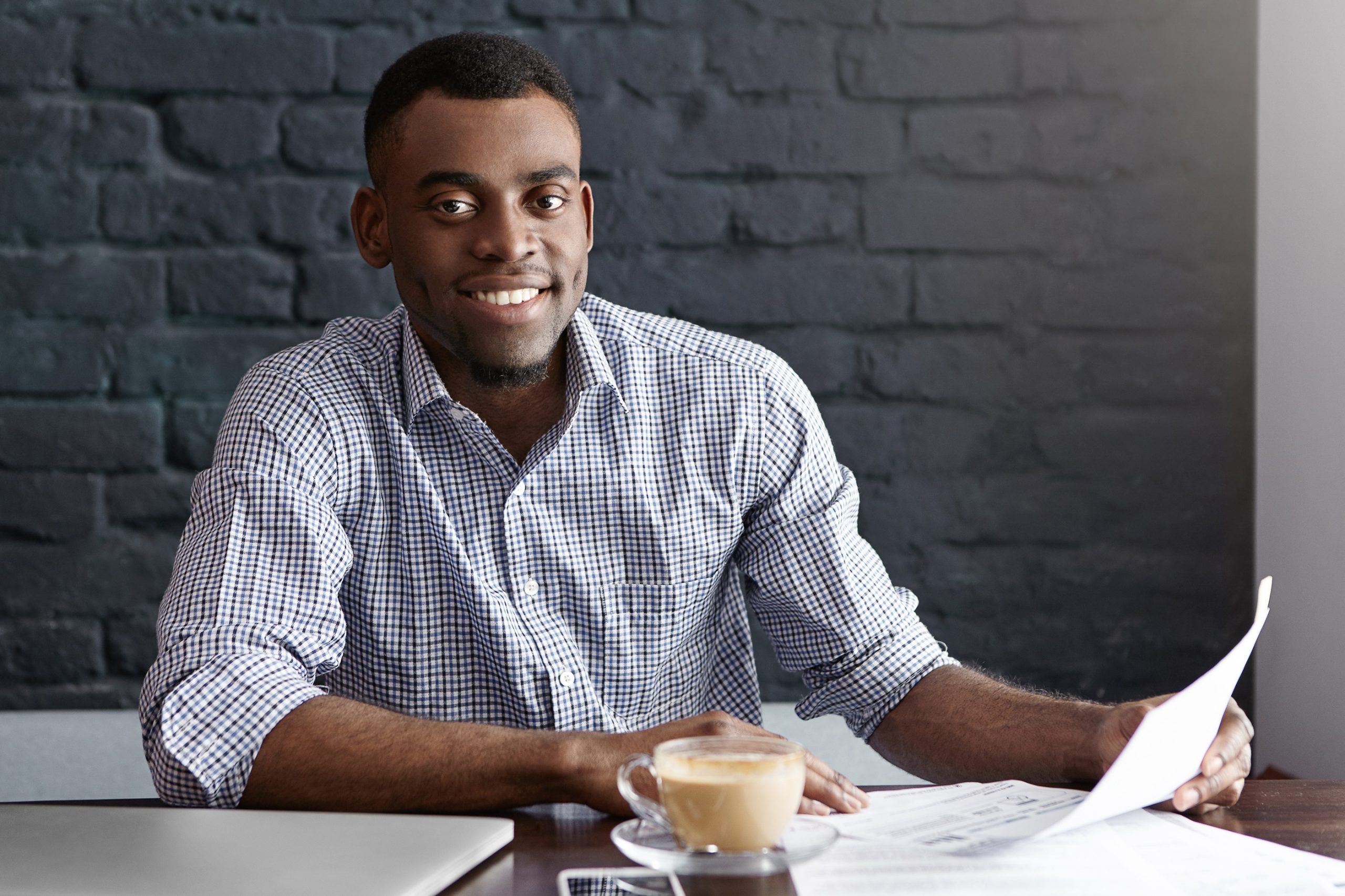 Cheerful successful young African entrepreneur holding piece of paper, happy with good numbers in financial report while looking through documents at desk in his office. People and business concept Cheerful successful young African entrepreneur holding piece of paper, happy with good numbers in financial report while looking through documents at desk in his office. People and business concept
