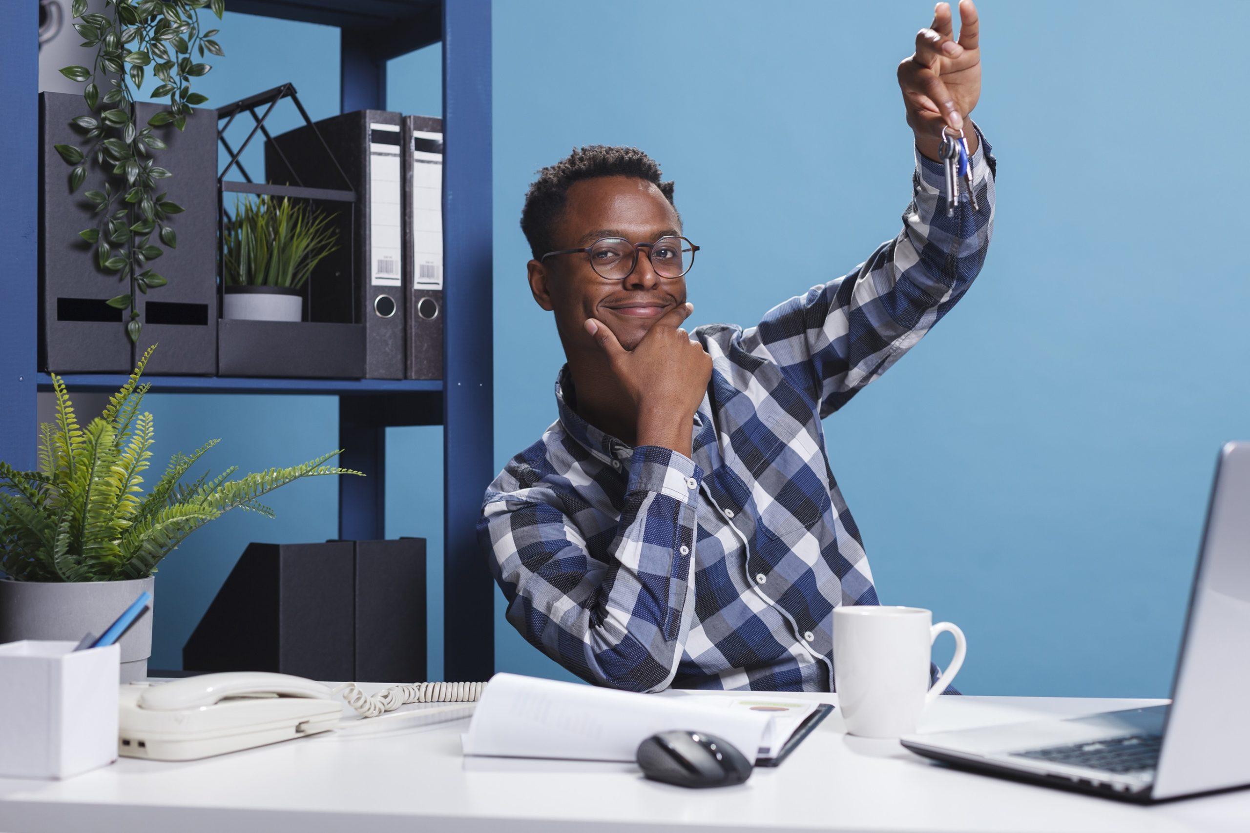 Young adult home owner holding keys of newly bought house while in office. Young adult home owner holding keys of new bought house while in office. Confident and happy man having keys to lately estate property investment while sitting at desk in modern workspace.